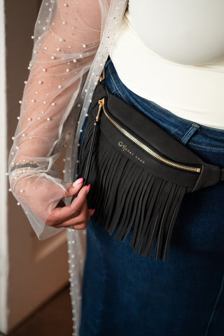 Person wearing a black fringed waist bag with a visible brand name.