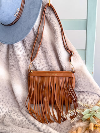 Brown fringe handbag with 'Gray Malin' branding on a textured surface with a blue hat and plant.