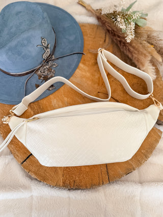 White crossbody bag on a wooden surface with a blue hat and dried flowers in the background