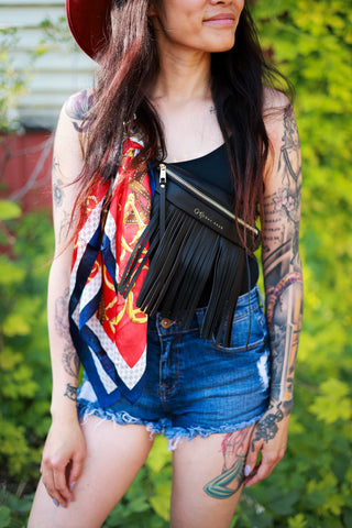 Person wearing a colorful scarf, black fringe bag, and denim shorts with tattoos on arms and legs.