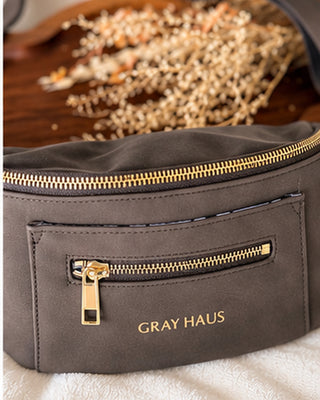 A dark gray faux-leather waist bag with gold-tone zippers and the brand name 'GRAY HAUS' printed in gold on the front pocket, resting on a white textured surface with dried flowers in the background.
