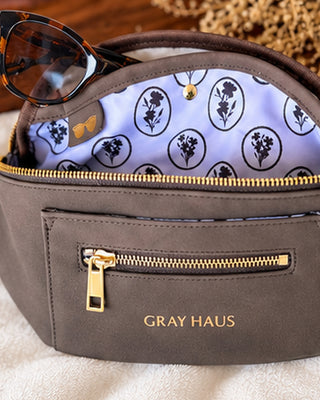 Gray Haus cosmetic pouch in taupe brown with gold hardware, featuring a front zip pocket and an open top revealing a lavender interior with black floral motifs. Sunglasses rest inside, and the brand name 'GRAY HAUS' is printed in gold on the front.