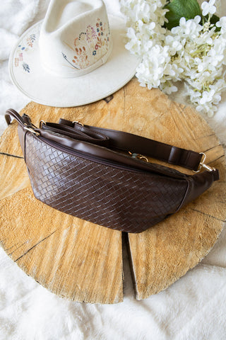 Brown woven belt bag on a wooden surface with a white flower arrangement and ceramic items.
