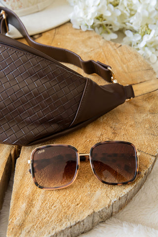 Brown woven handbag and sunglasses on a wooden surface with white flowers in the background