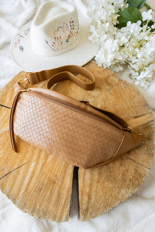 Brown woven handbag on a wooden surface with a white hat and flowers in the background