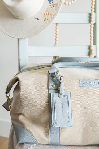 Beige duffel bag with blue accents and a matching tag on a chair with a hat and decorative elements.