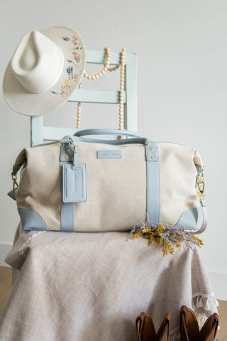 Beige handbag with blue accents on a chair with a hat, shoes, and flowers.