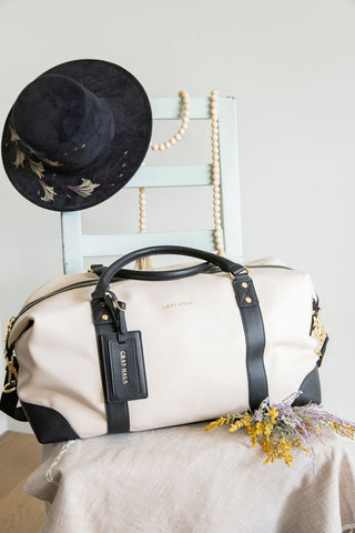 Beige duffel bag with black straps on a chair next to a black hat and decorative items.