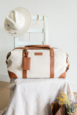 White duffel bag with brown leather straps on a white surface with a hat and flowers in the background.
