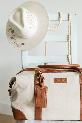 Canvas duffel bag with brown leather accents and a cowboy hat on a wooden surface.