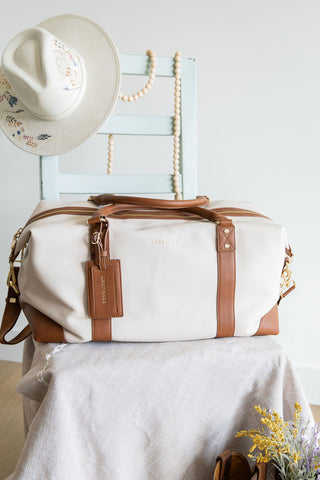 White duffel bag with brown leather straps on a white surface, with a white hat and decorative elements in the background.