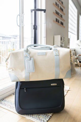 Two bags, a beige duffel bag and a black suitcase, on a wooden floor with a window in the background.