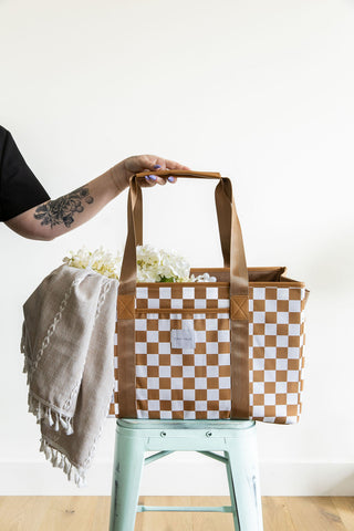 Person holding a checkered bag with flowers and a blanket on a stool against a white background