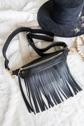 Black fringe handbag with gold accents on a white surface with a black hat and wooden stick in the background.