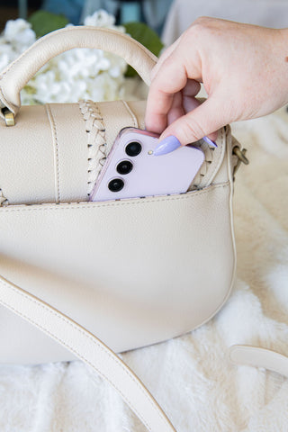 Hand placing a smartphone into a beige handbag on a white surface