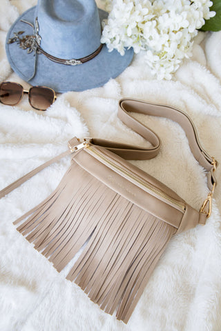 Beige fringe handbag with sunglasses and a blue hat on a white surface