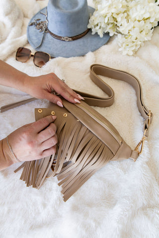 Beige fringe handbag held by a hand on a white fluffy surface with a hat, sunglasses, and flowers in the background.