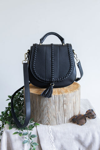 Black handbag with tassels on a wooden block with sunglasses and plant in the background