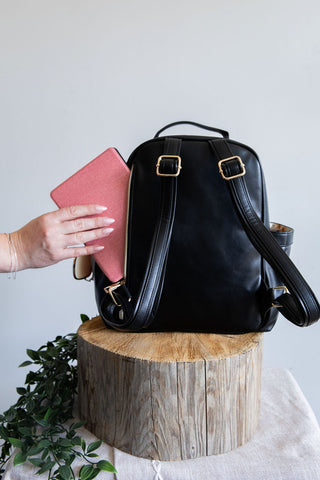 Black leather backpack on a wooden stump with a pink book being placed into it.