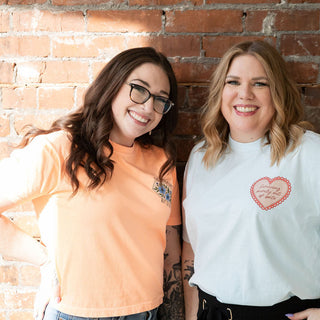 Two women standing against a brick wall, one wearing an orange shirt and glasses, the other in a white shirt with a heart design.